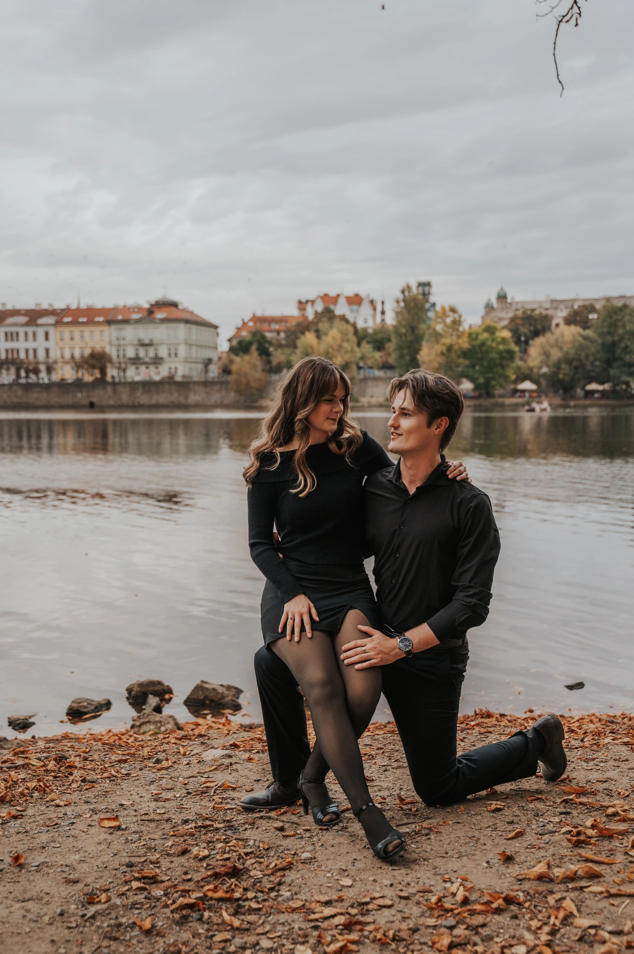 Couple sharing a playful moment on the leaf-covered riverbank during a couple photoshoot in Prague.