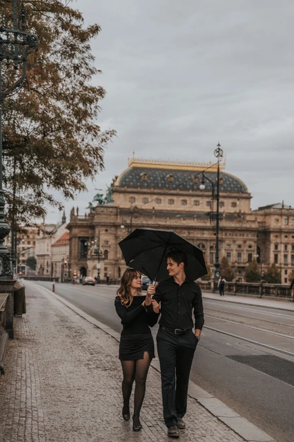 Prague couple photoshoot: couple walking under an umbrella with the National Theatre behind them. Couple walking under an umbrella with the National Theatre behind them during a couple photoshoot in Prague.