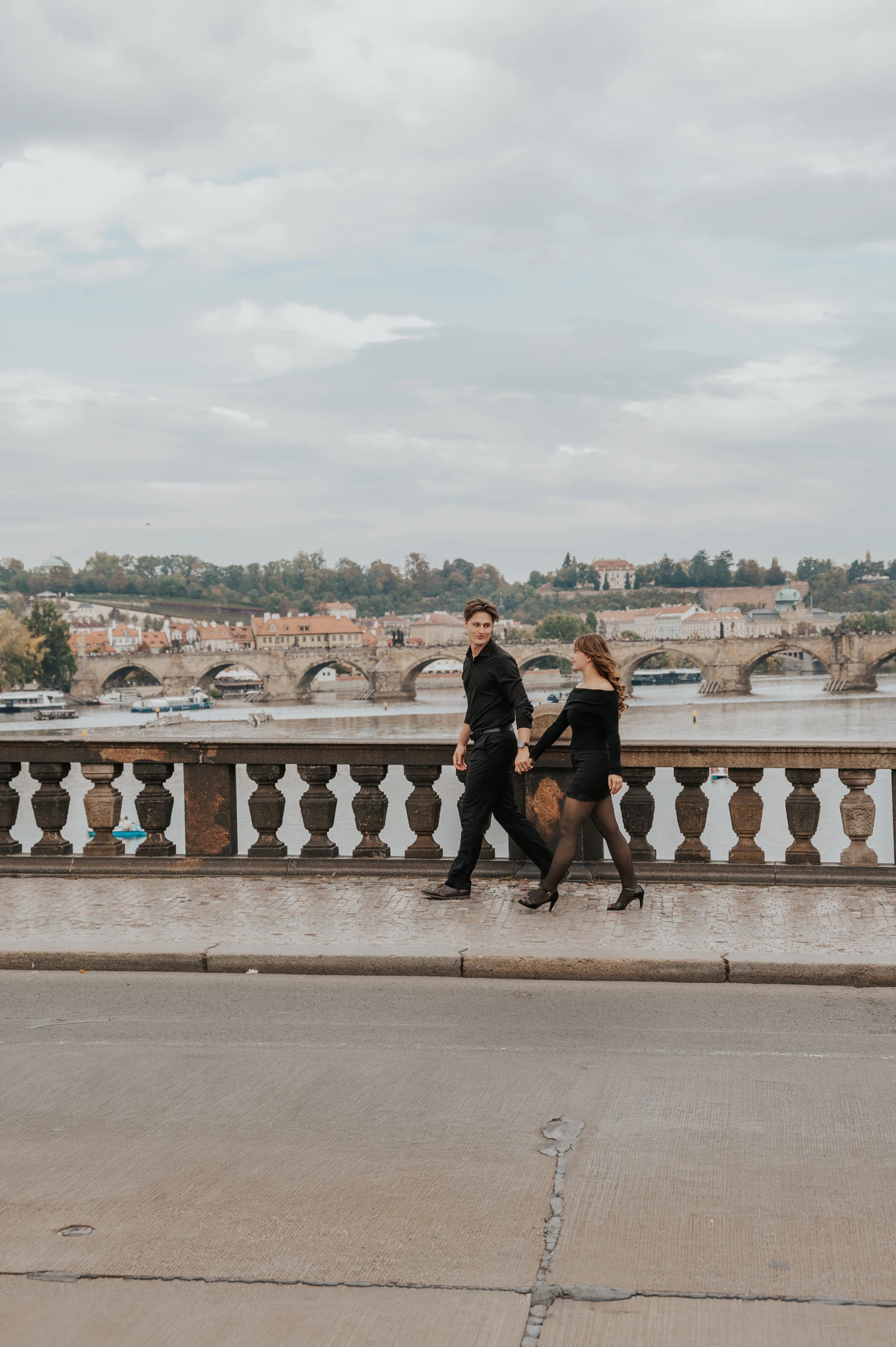 Wide view of the couple walking along the Legion Bridge balustrade during a couple photoshoot in Prague.