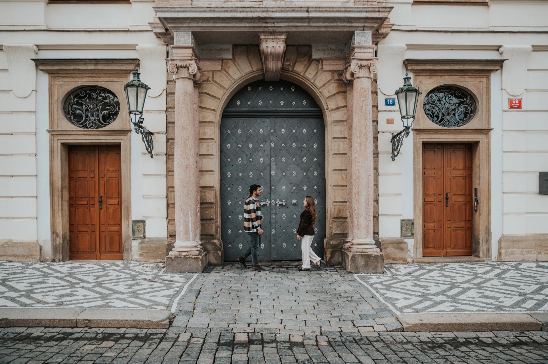 Wide photograph of the couple standing apart beneath a grand arched gateway with wooden doors on either side during their Prague couple photoshoot.