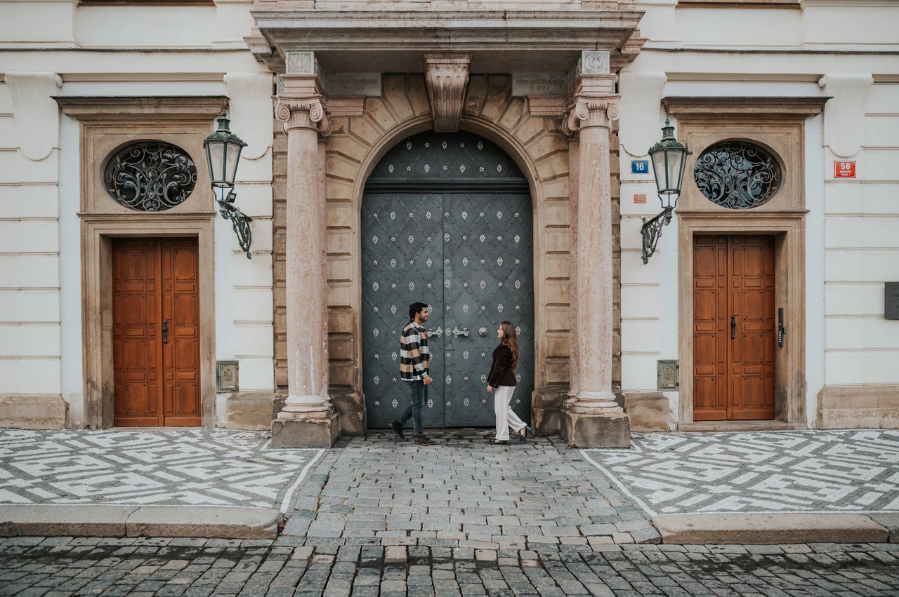 Prague couple photoshoot: a wide view of the ornate castle gateway Wide photograph of the couple standing apart beneath a grand arched gateway with wooden doors on either side during their Prague couple photoshoot.