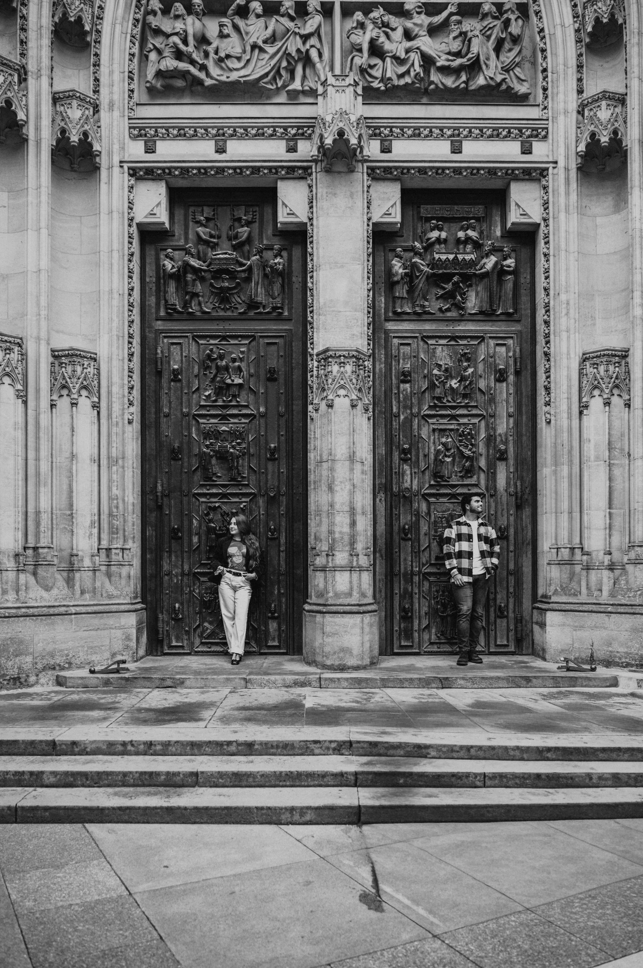 Black-and-white photograph of the couple standing apart in front of ornate cathedral doors during their Prague couple photoshoot.