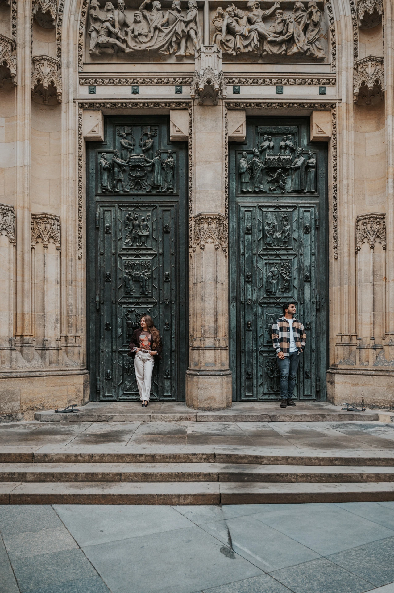 Couple standing on either side of a stone column beneath tall bronze cathedral doors during their Prague couple photoshoot.
