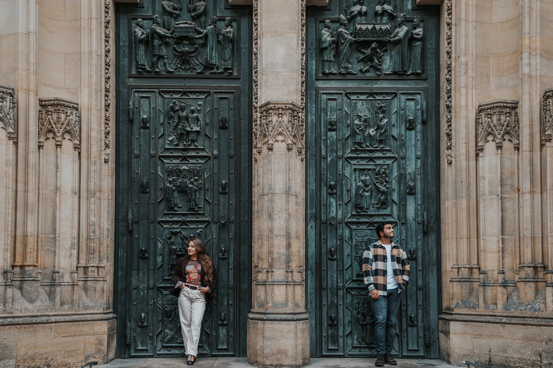 Wide view of the couple on the cathedral steps beneath carved bronze doors during their Prague couple photoshoot.
