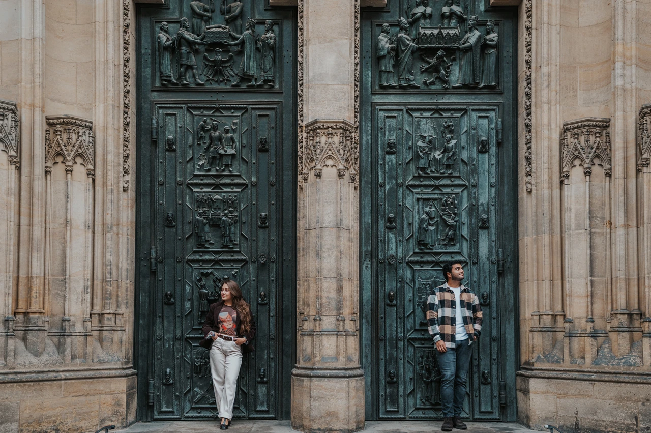 Prague couple photoshoot: a wide portrait beneath the bronze cathedral doors Wide view of the couple on the cathedral steps beneath carved bronze doors during their Prague couple photoshoot.