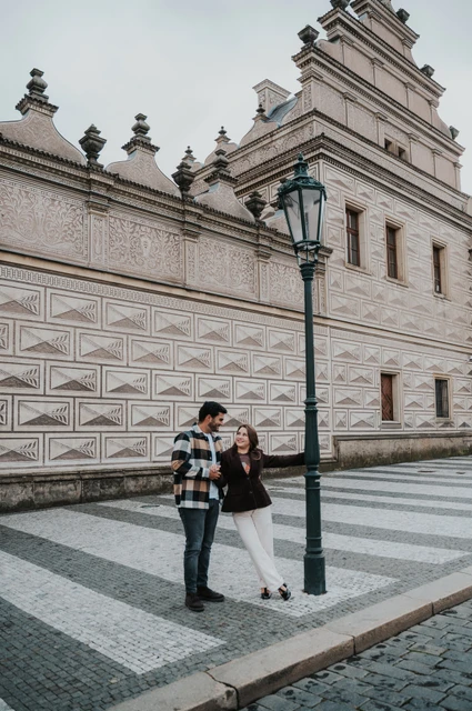 Prague couple photoshoot: hand in hand on the castle square Couple standing hand in hand on patterned paving beside a lamppost and a decorated facade during their Prague couple photoshoot.