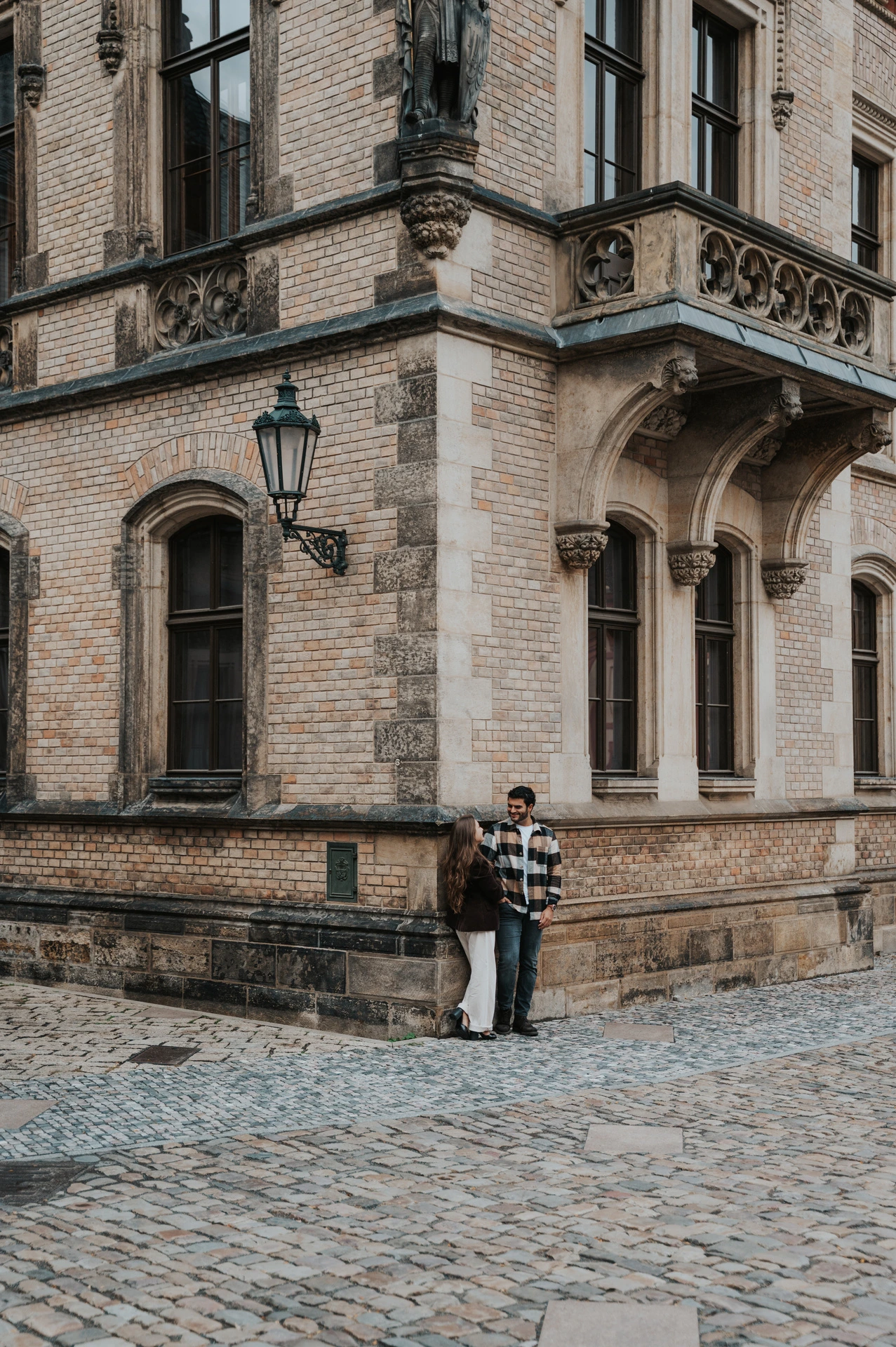 Distant portrait of the couple together at a stone corner beneath tall cathedral windows during their Prague couple photoshoot.