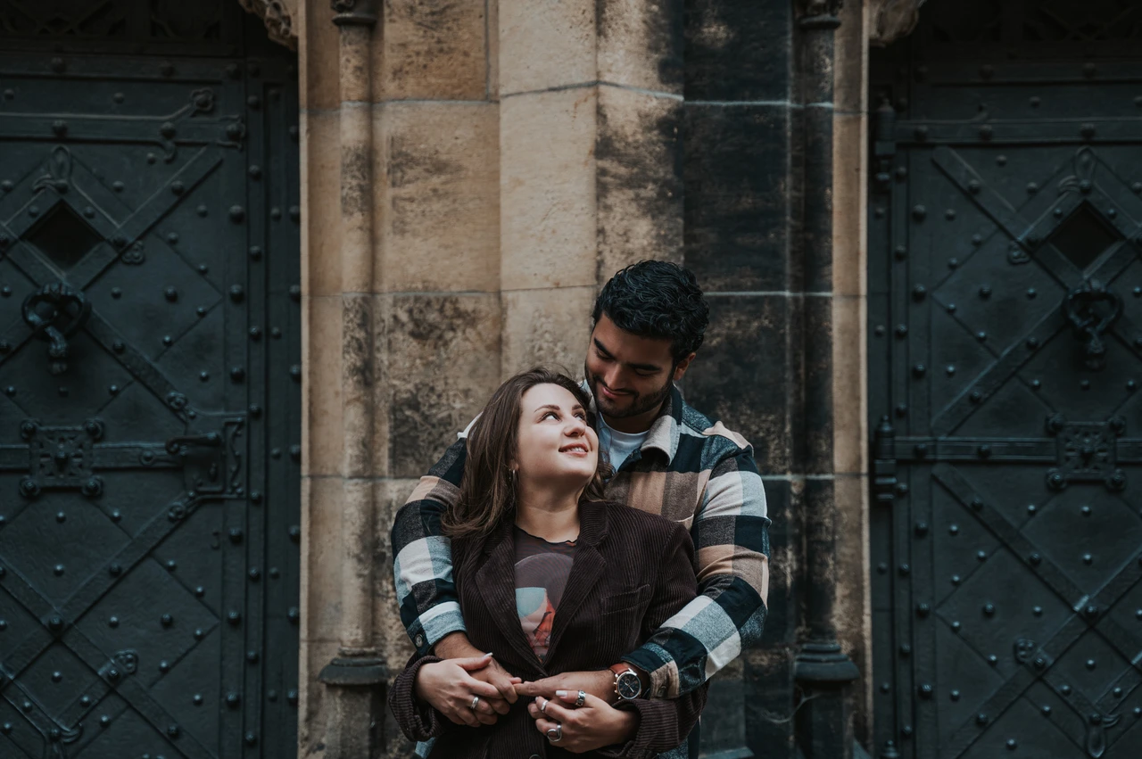 Prague couple photoshoot: a close embrace beside the cathedral doors Man embracing his partner from behind as they smile beside dark cathedral doors during their Prague couple photoshoot.