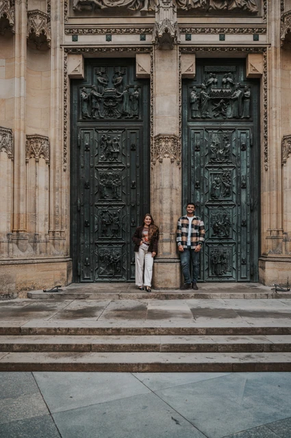 Prague couple photoshoot: with Amigo by the carved cathedral doors Couple standing apart beneath tall carved cathedral doors, with the woman holding their dog Amigo during their Prague couple photoshoot.