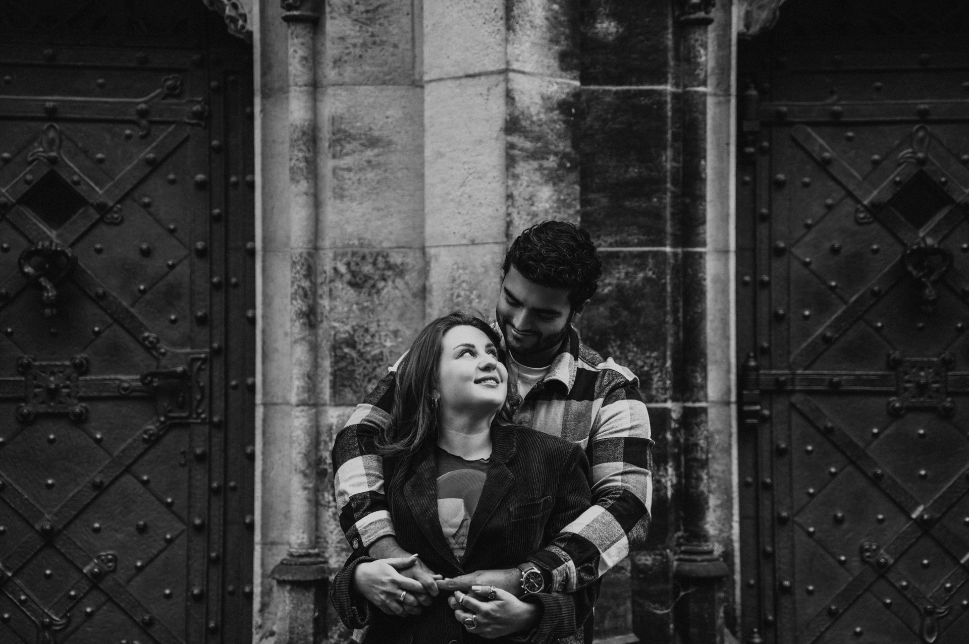 Black-and-white landscape portrait of the couple smiling in an embrace by the cathedral doors during their Prague couple photoshoot.