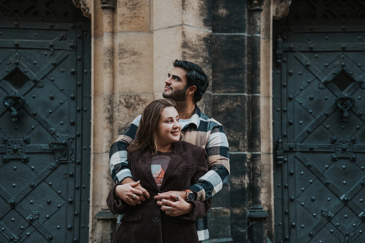 Prague couple photoshoot: an embrace among the cathedral's stone details Wide portrait of the couple looking upwards while embracing beside carved stone and dark doors during their Prague couple photoshoot.