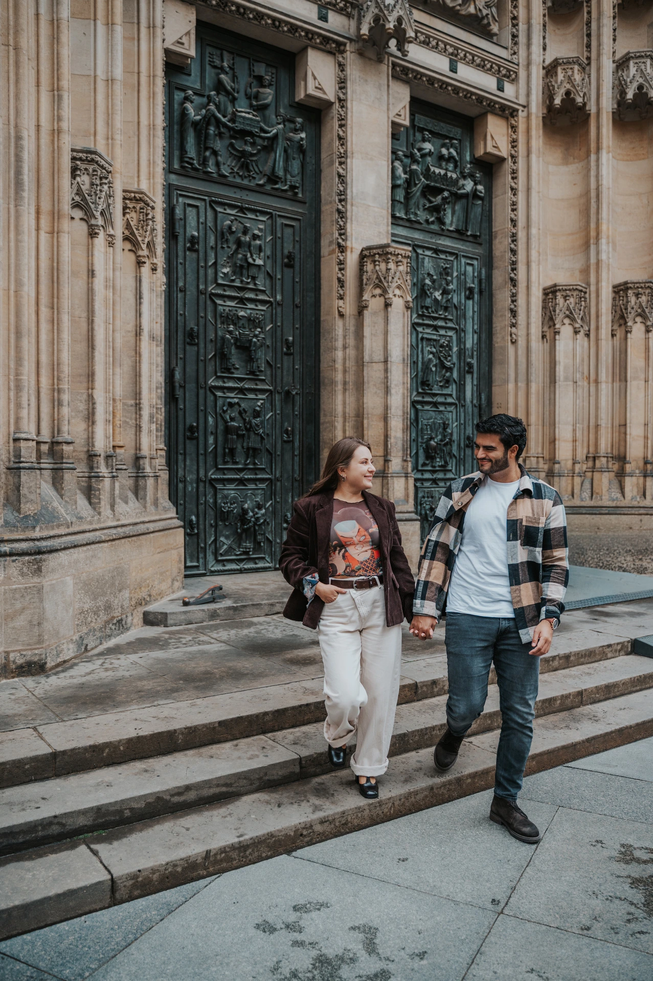 Couple walking down the cathedral steps while the woman carries their dog Amigo during their Prague couple photoshoot.