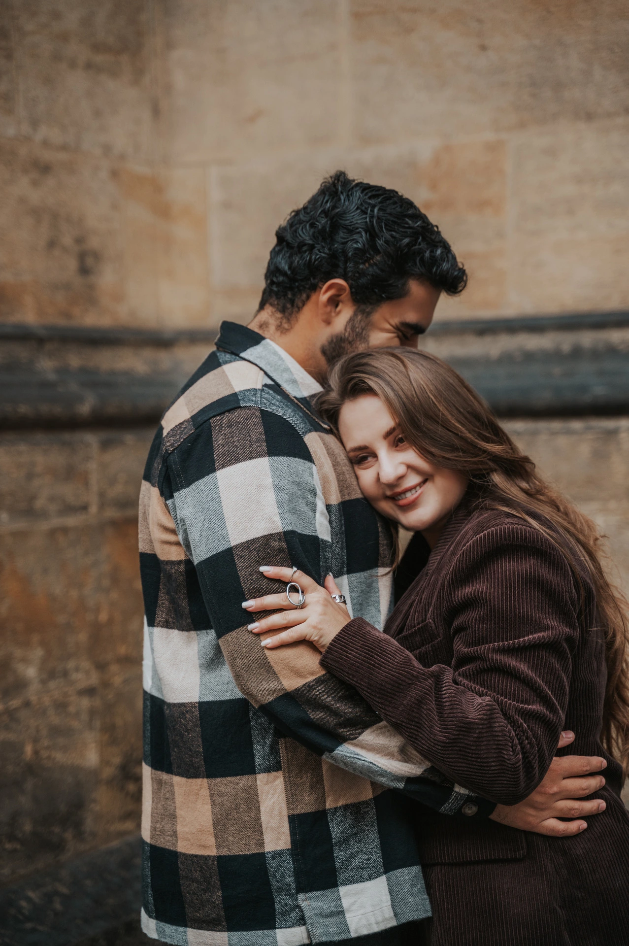 Woman resting against her partner's chest as he embraces her beside a stone wall during their Prague couple photoshoot.