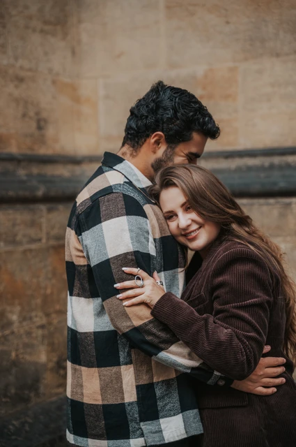 Prague couple photoshoot: a close embrace beside the castle stonework Woman resting against her partner's chest as he embraces her beside a stone wall during their Prague couple photoshoot.