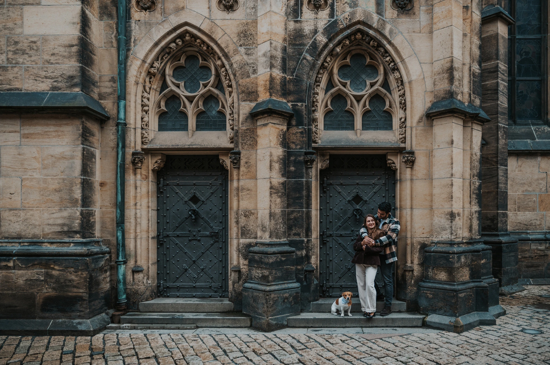Wide photograph of the couple walking in front of the cathedral's pointed Gothic doorways during their Prague couple photoshoot.