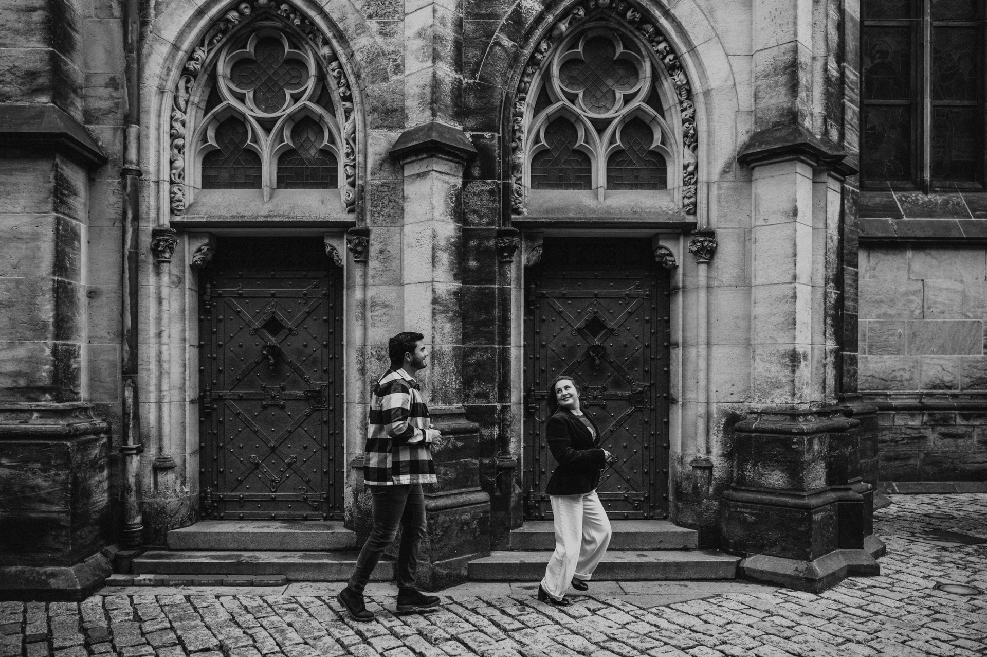 Black-and-white wide photograph of the couple walking past Gothic cathedral doorways during their Prague couple photoshoot.