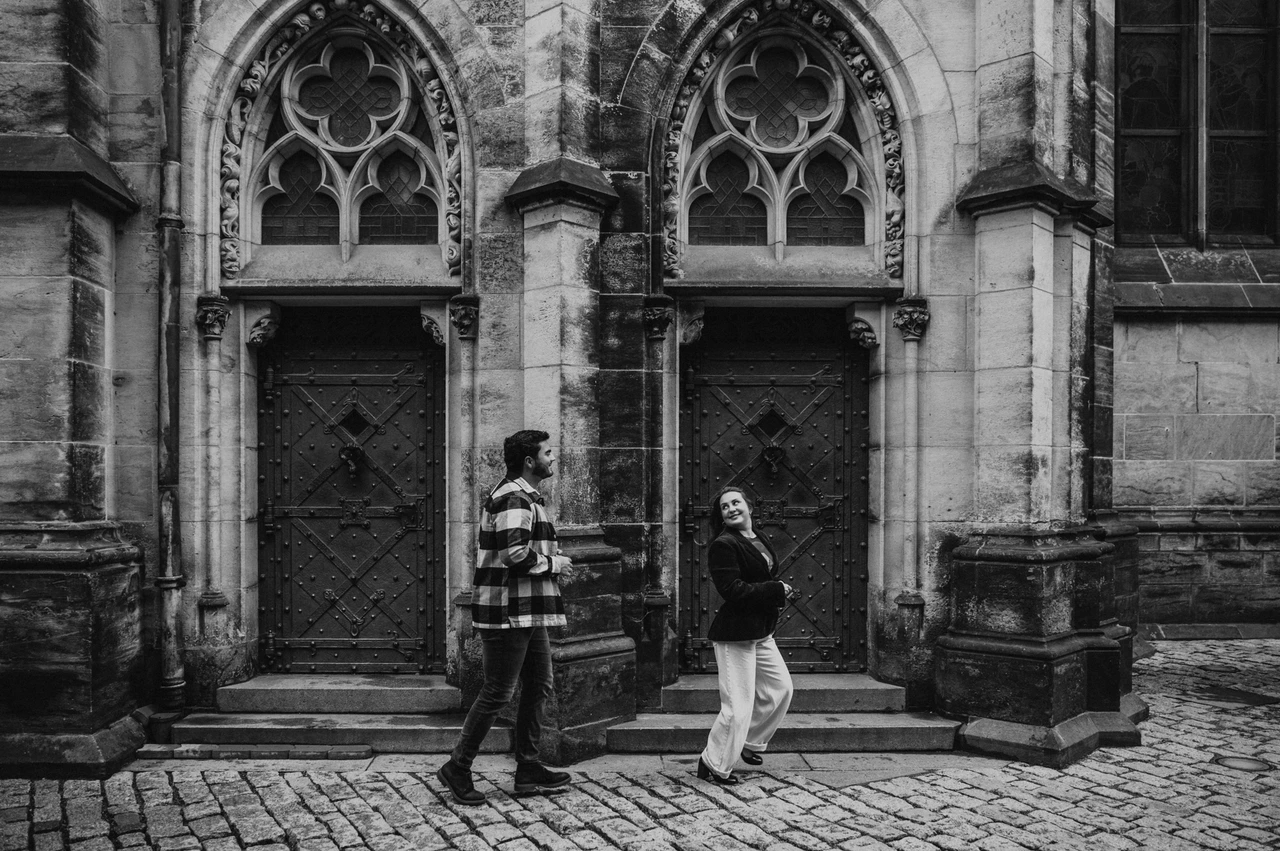 Prague couple photoshoot: a black-and-white walk beside the cathedral Black-and-white wide photograph of the couple walking past Gothic cathedral doorways during their Prague couple photoshoot.