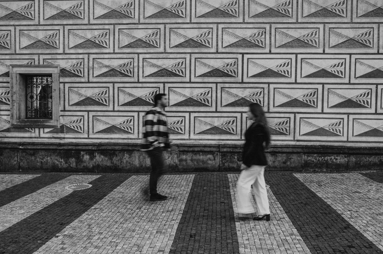 Prague couple photoshoot: a black-and-white walk across the castle square Black-and-white wide photograph of the couple walking towards each other beside a geometric facade during their Prague couple photoshoot.