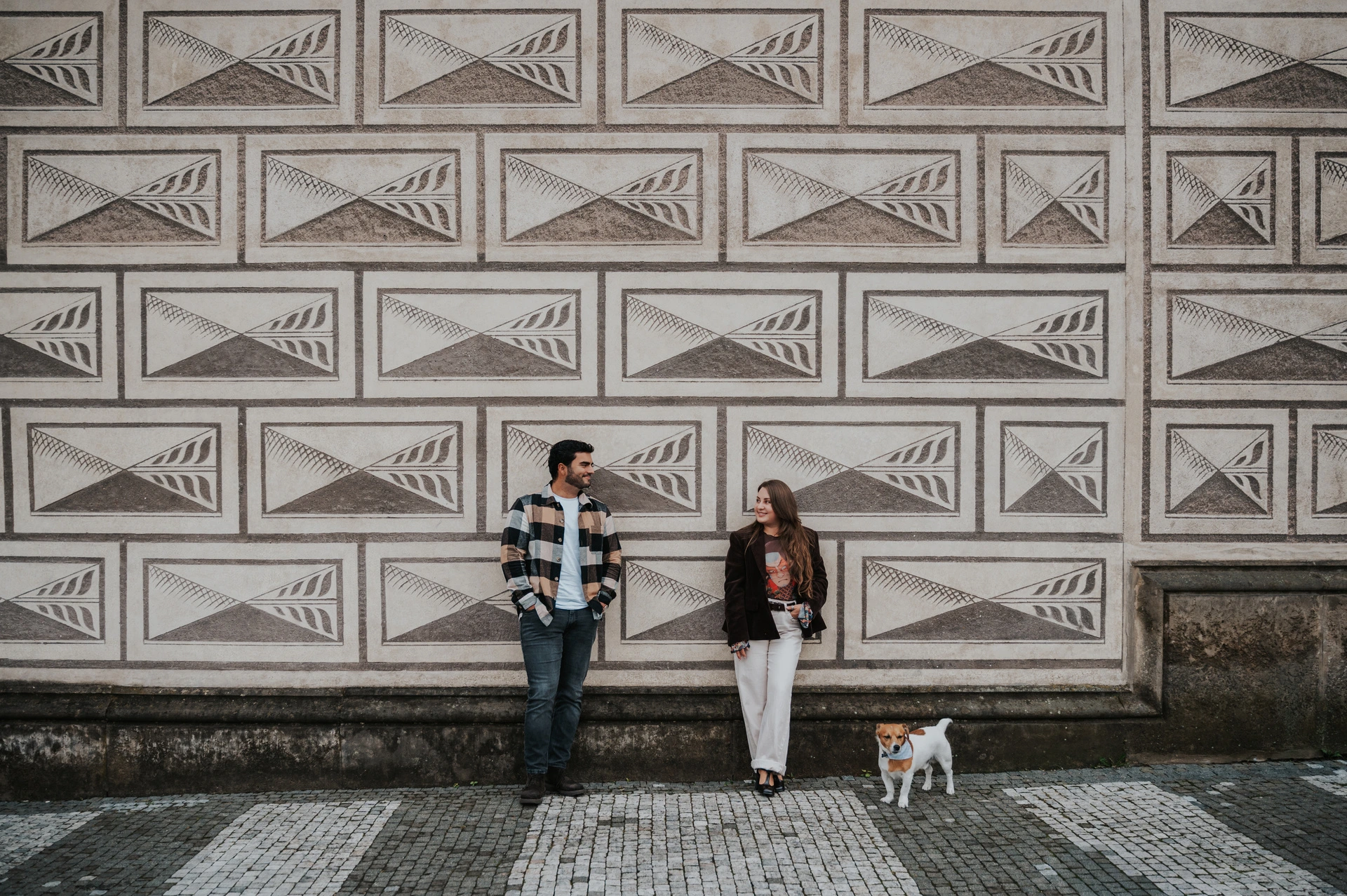 Couple standing apart beside a patterned stone facade, with their dog Amigo between them during their Prague couple photoshoot.