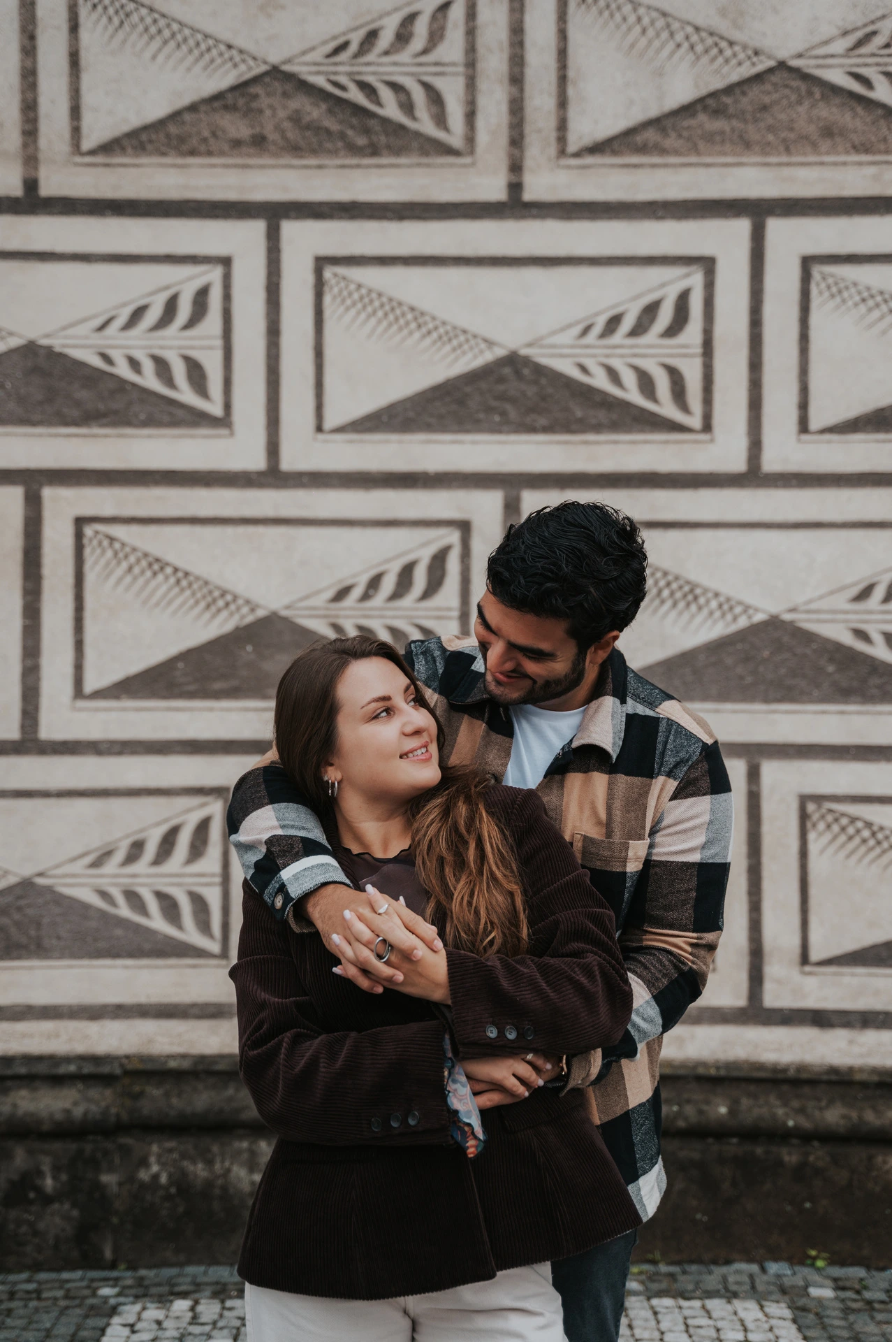 Man embracing his smiling partner from behind beside a patterned stone wall during their Prague couple photoshoot.