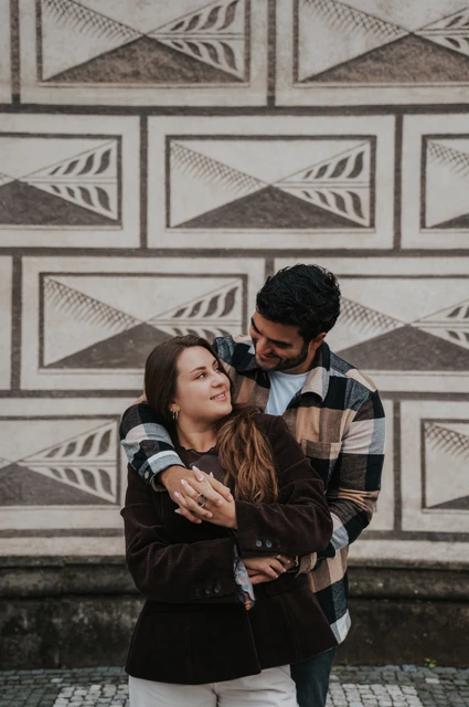Prague couple photoshoot: a close embrace by the patterned facade Man embracing his smiling partner from behind beside a patterned stone wall during their Prague couple photoshoot.
