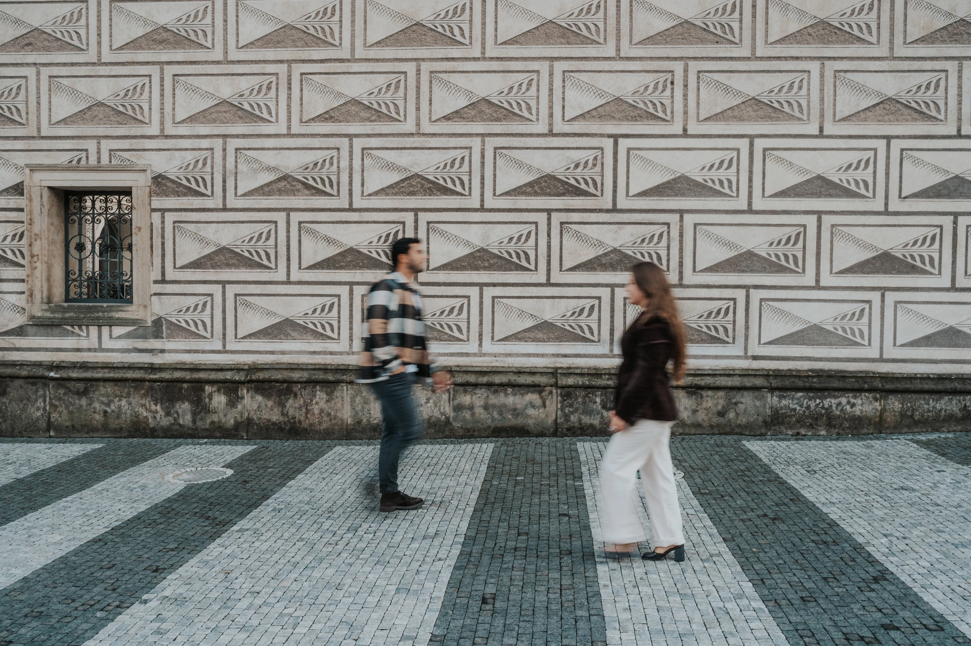 Couple walking towards each other across patterned paving beside a geometric stone facade during their Prague couple photoshoot.