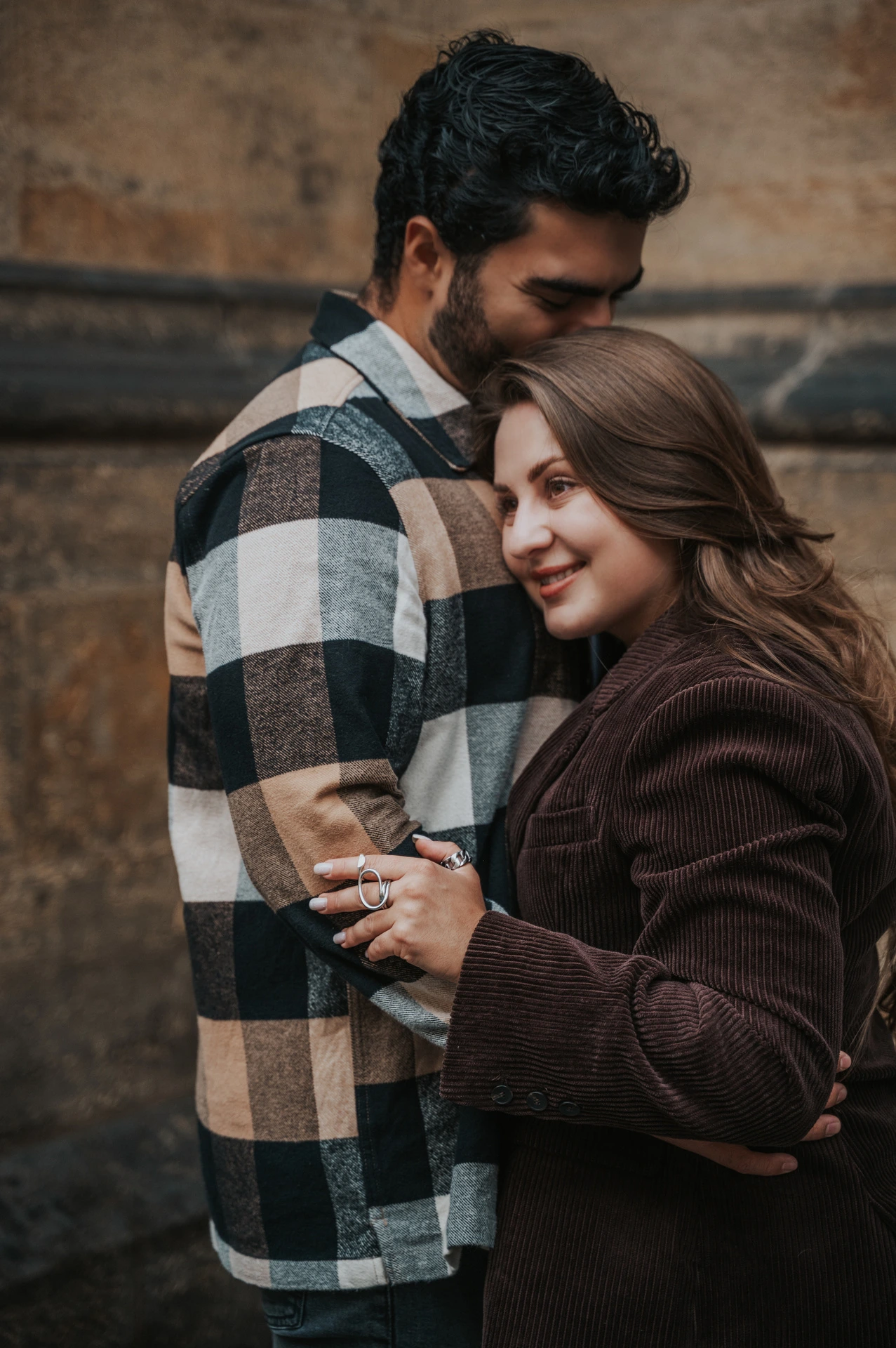 Woman smiling towards the camera while leaning against her partner beside a stone wall during their Prague couple photoshoot.