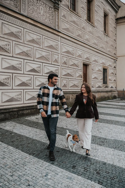 Couple walking beside a patterned facade with their small dog Amigo on a lead during their Prague couple photoshoot.