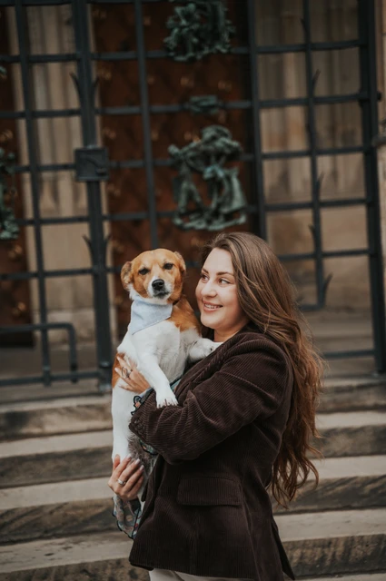 Woman holding her small dog Amigo in front of a decorated cathedral door during their Prague couple photoshoot.
