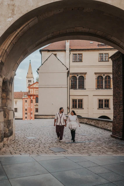 Prague couple photoshoot beneath a castle archway Couple walking hand in hand beneath a stone archway, with the castle courtyard behind them, during their Prague photoshoot.
