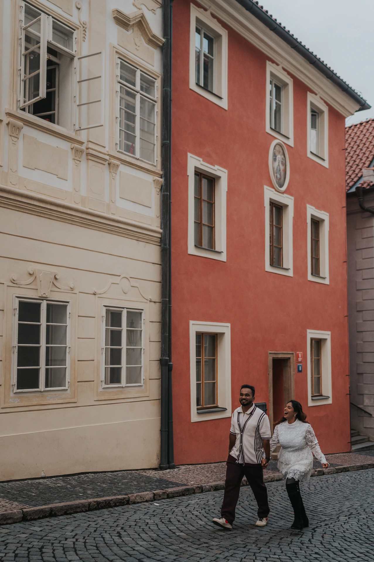 Couple walking hand in hand along a cobbled street beside red and cream houses during their Prague Castle photoshoot.