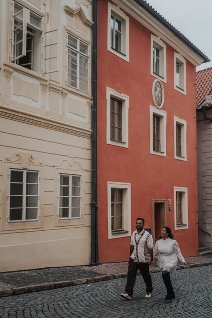 Prague photoshoot on a colourful street below the castle Couple walking hand in hand along a cobbled street beside red and cream houses during their Prague Castle photoshoot.