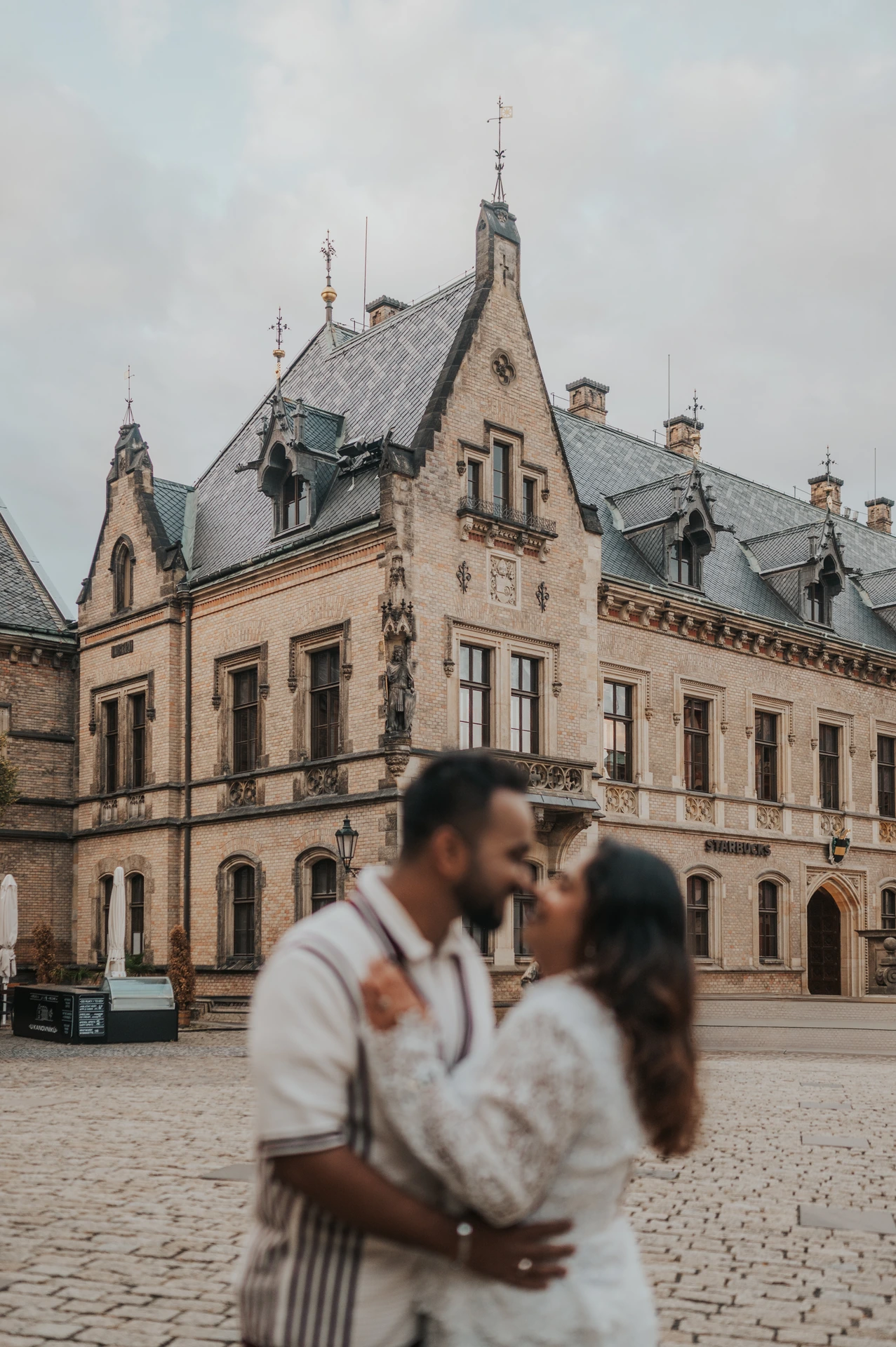 Soft-focus couple kissing in the foreground with the castle courtyard architecture in focus during their Prague photoshoot.