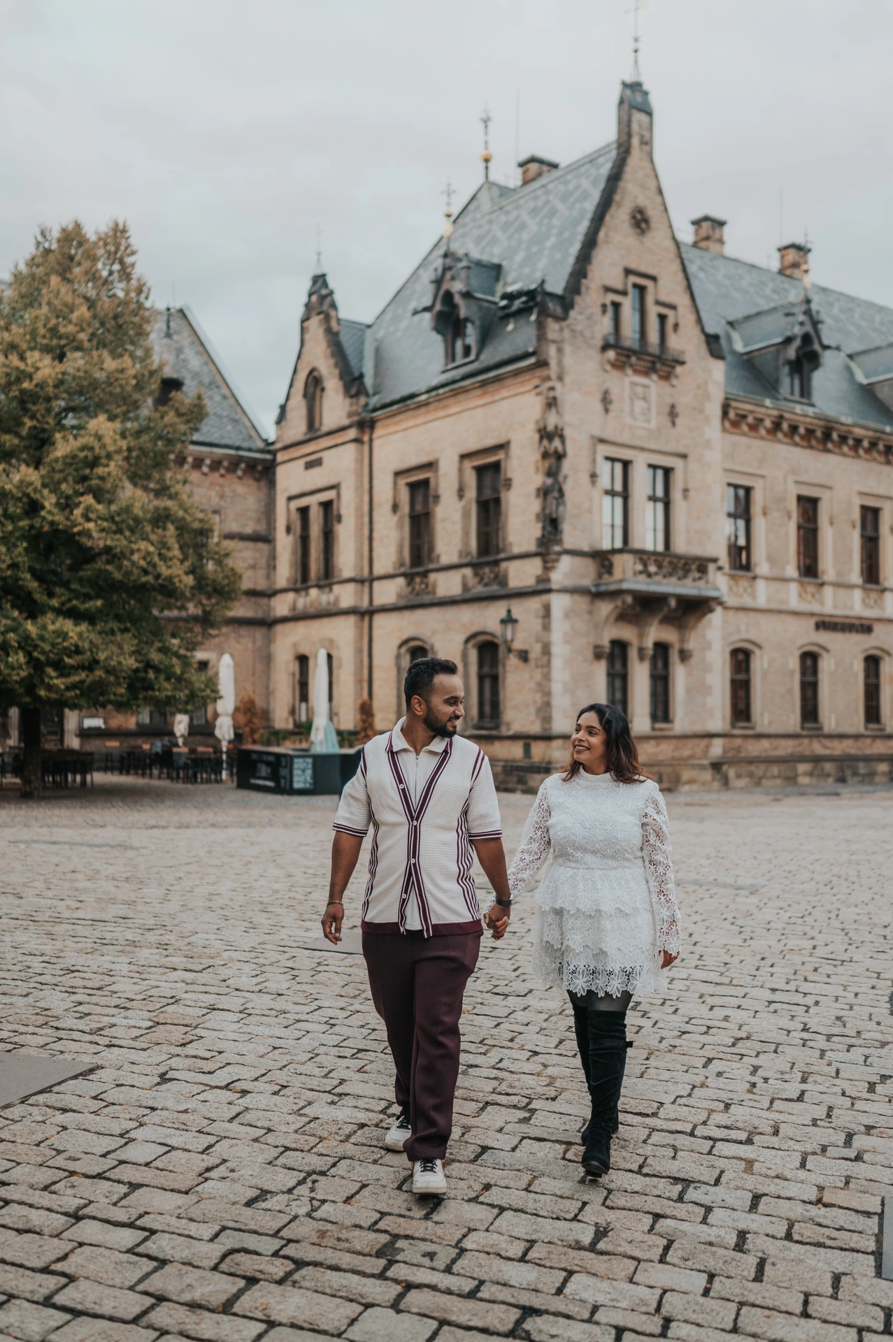 Couple walking hand in hand across a cobbled castle courtyard with an ornate building behind them during their Prague photoshoot.