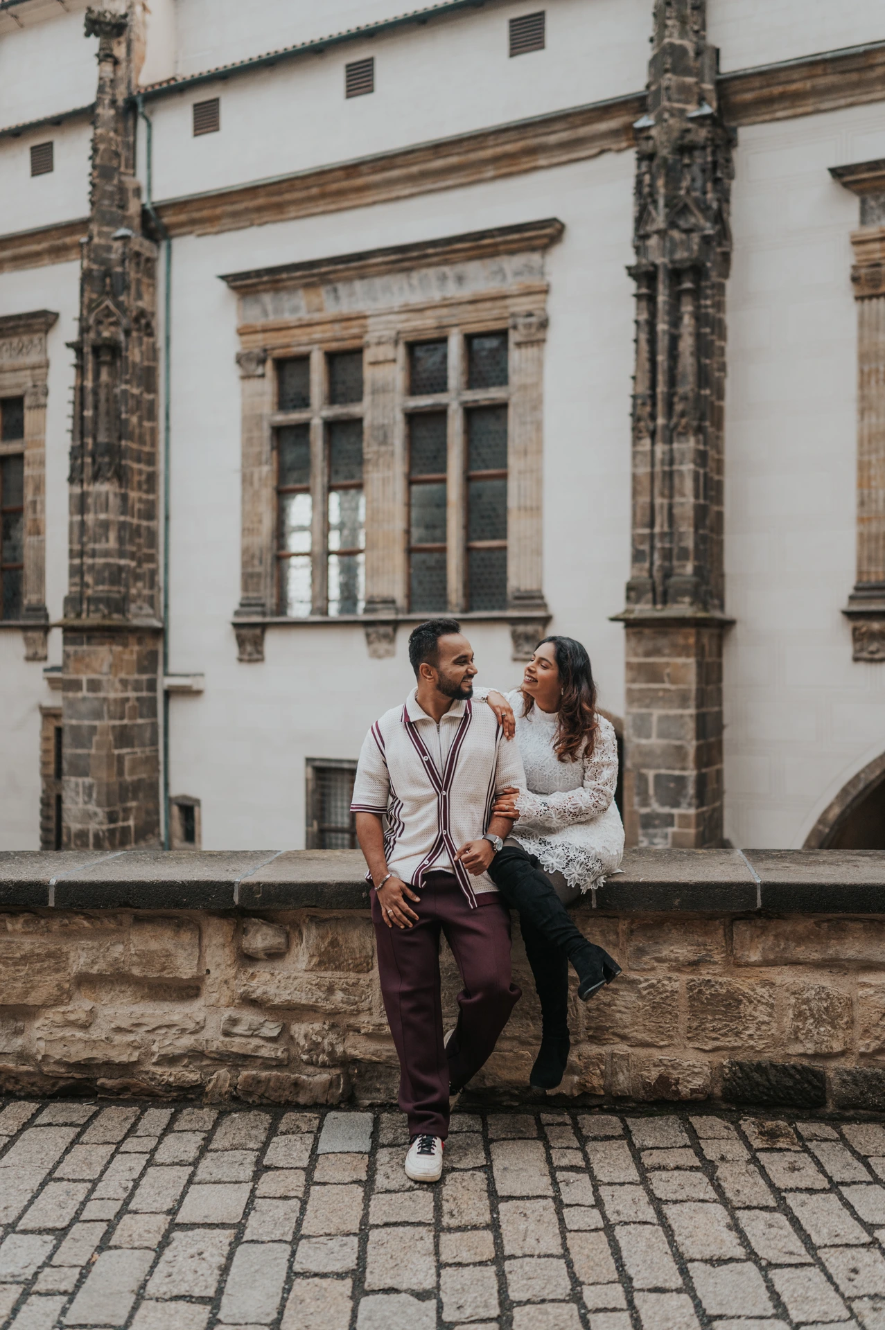 Couple sitting together on a stone ledge beside the castle facade during their couple photoshoot in Prague.