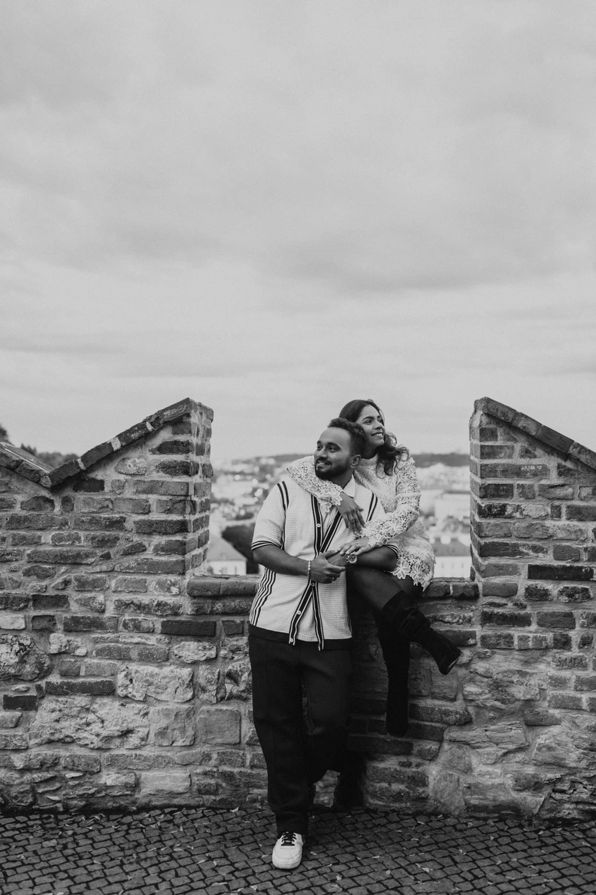 Prague couple photoshoot with a playful embrace at the castle viewpoint Black-and-white portrait of the man laughing as his wife embraces him from behind beside a stone viewpoint wall during their Prague photoshoot.