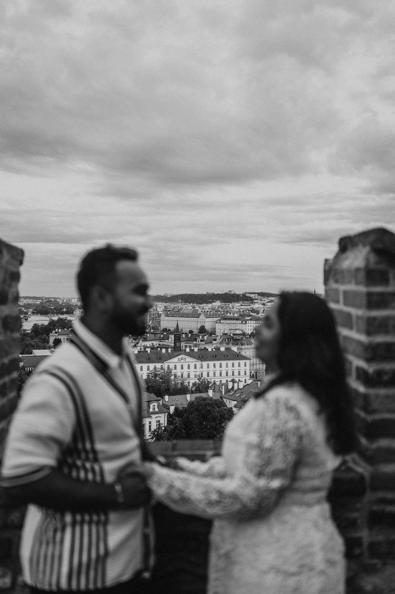 Black-and-white photograph with the couple softly blurred in the foreground and Prague rooftops beyond the viewpoint during their photoshoot.