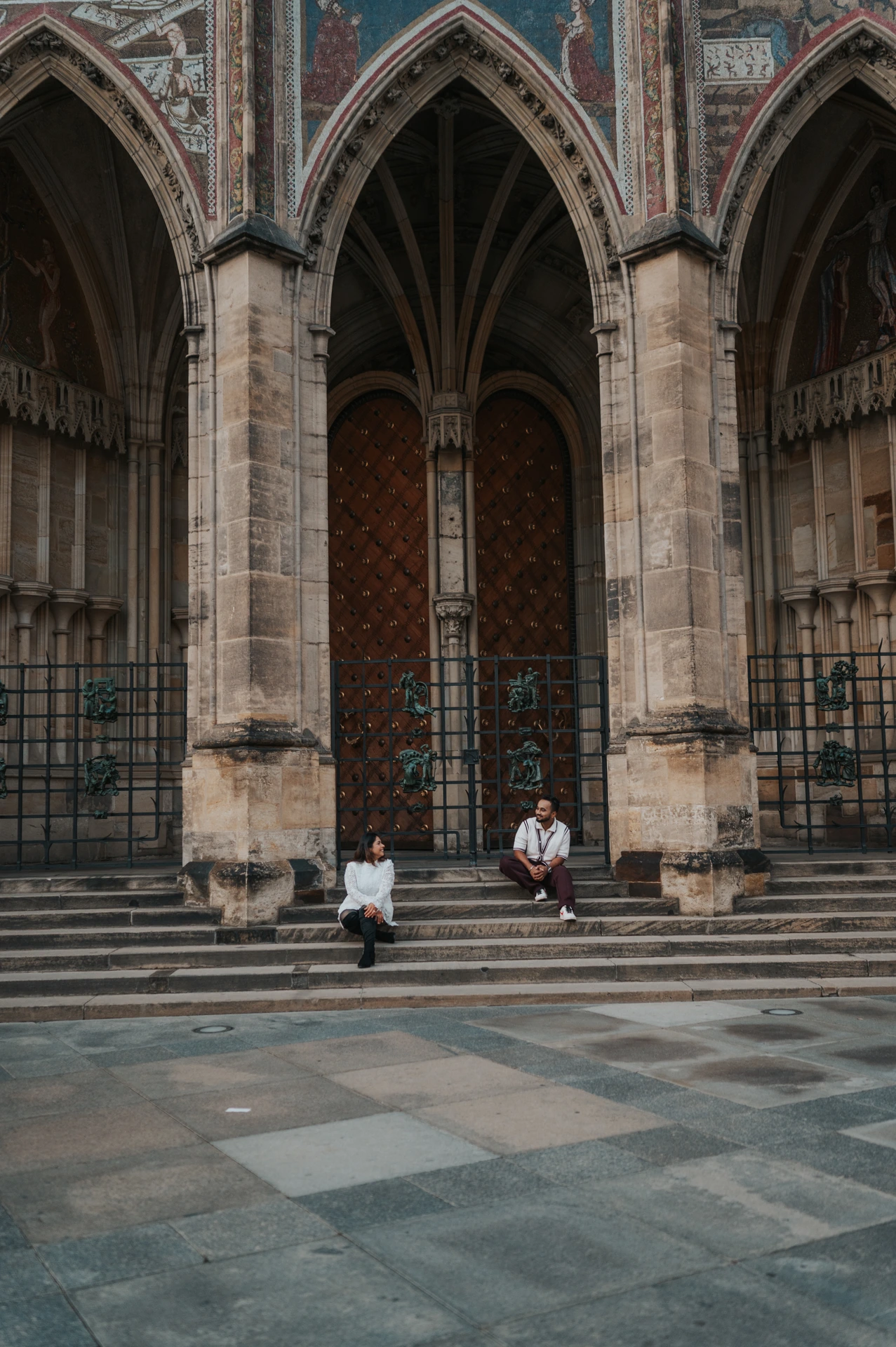 Wide vertical view of the couple seated apart beneath the pointed cathedral arches during their couple photoshoot at Prague Castle.