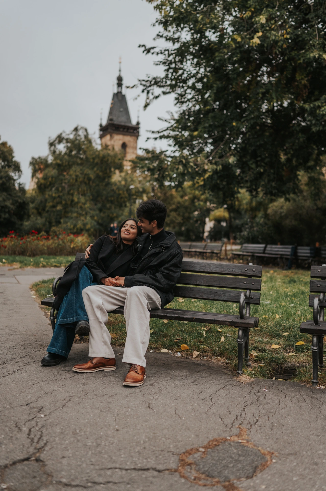 Couple embracing on a park bench beside fallen leaves and flowerbeds during their Prague couple photoshoot.