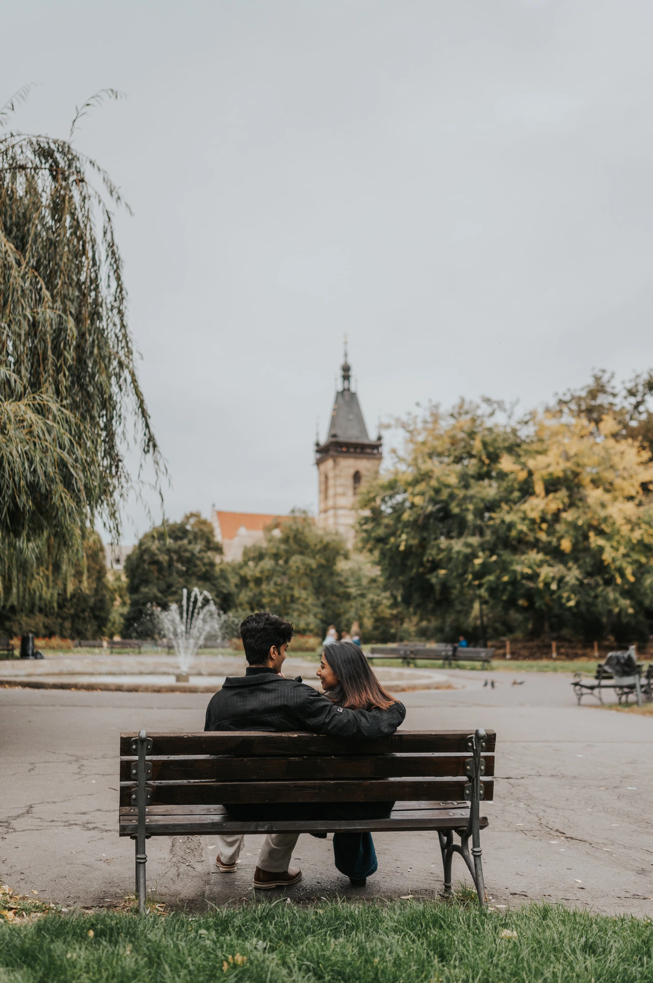 Rear view of the couple sitting close on a park bench facing the fountain and New Town Hall tower during their Prague couple photoshoot.