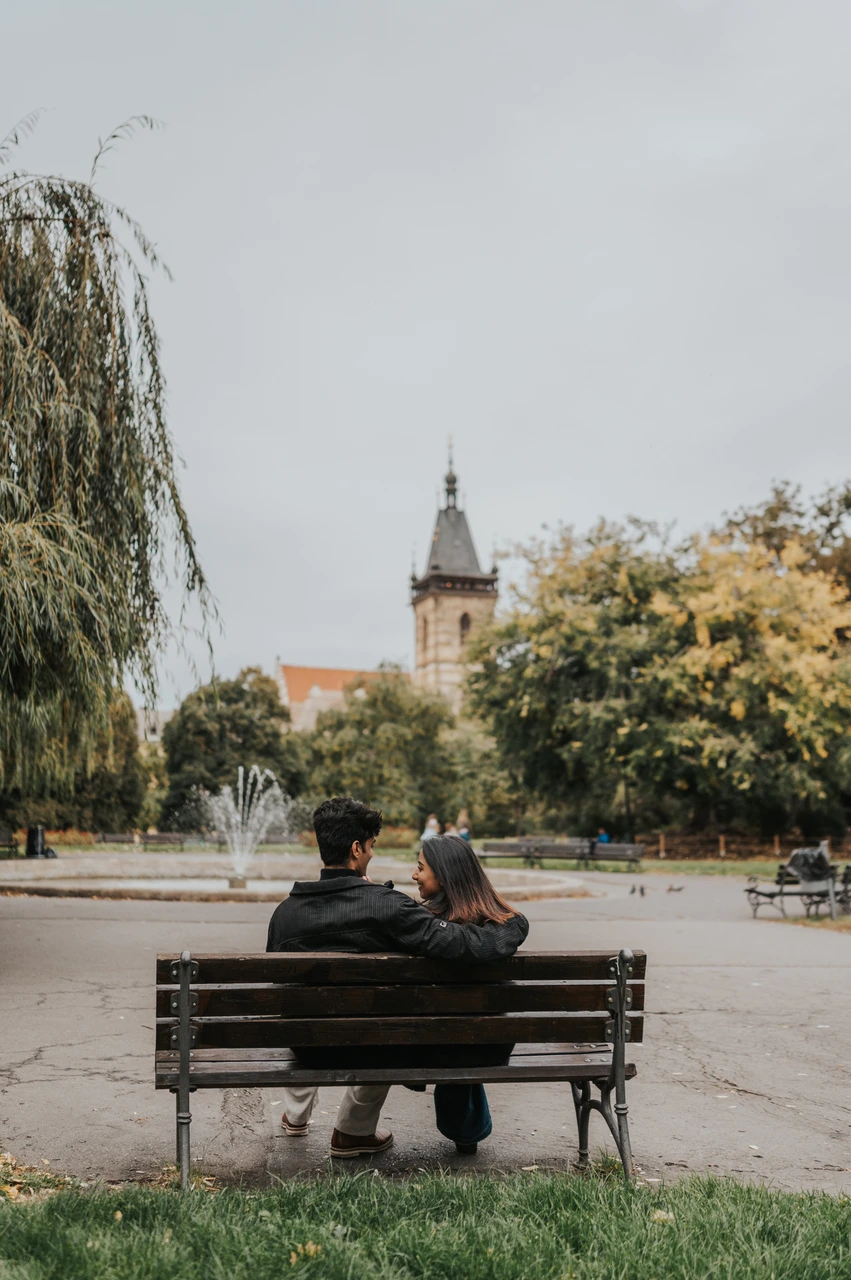 Prague couple photoshoot: a bench with a view of the fountain and tower Rear view of the couple sitting close on a park bench facing the fountain and New Town Hall tower during their Prague couple photoshoot.