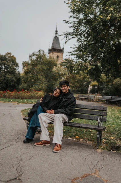 Prague couple photoshoot: smiles together on a Charles Square bench Couple sitting close together on a park bench and smiling with trees and the New Town Hall tower behind them during their Prague couple photoshoot.