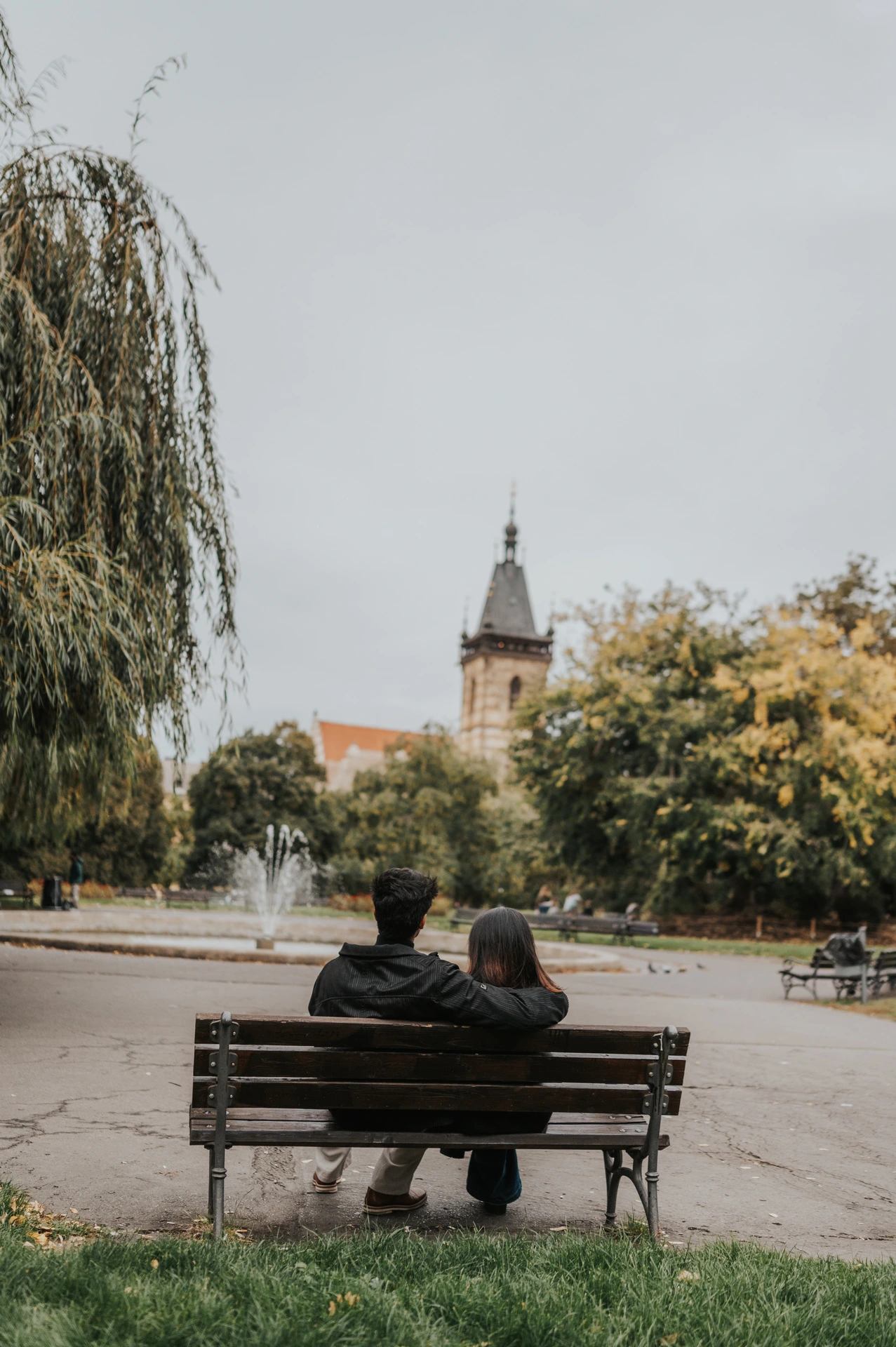 Couple seen from behind sharing a bench beneath the trees with the New Town Hall tower in the distance during their Prague couple photoshoot.