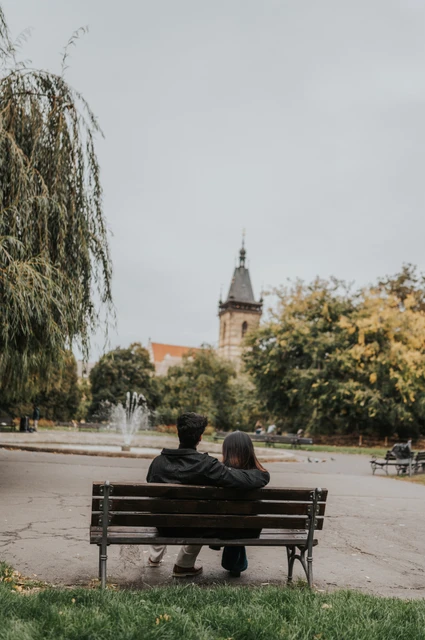 Prague couple photoshoot: a shared bench beneath the park trees Couple seen from behind sharing a bench beneath the trees with the New Town Hall tower in the distance during their Prague couple photoshoot.