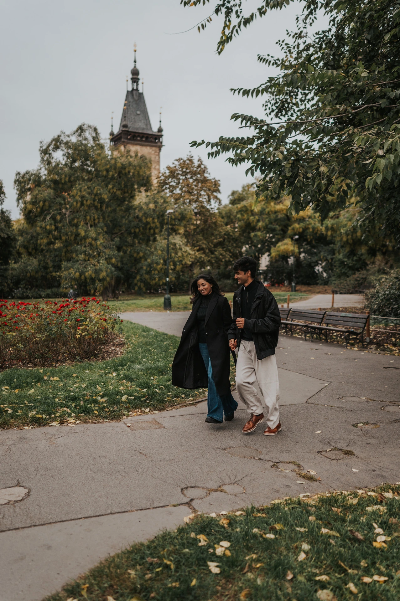 Couple walking hand in hand along a park path beside colourful flowerbeds during their Prague couple photoshoot.