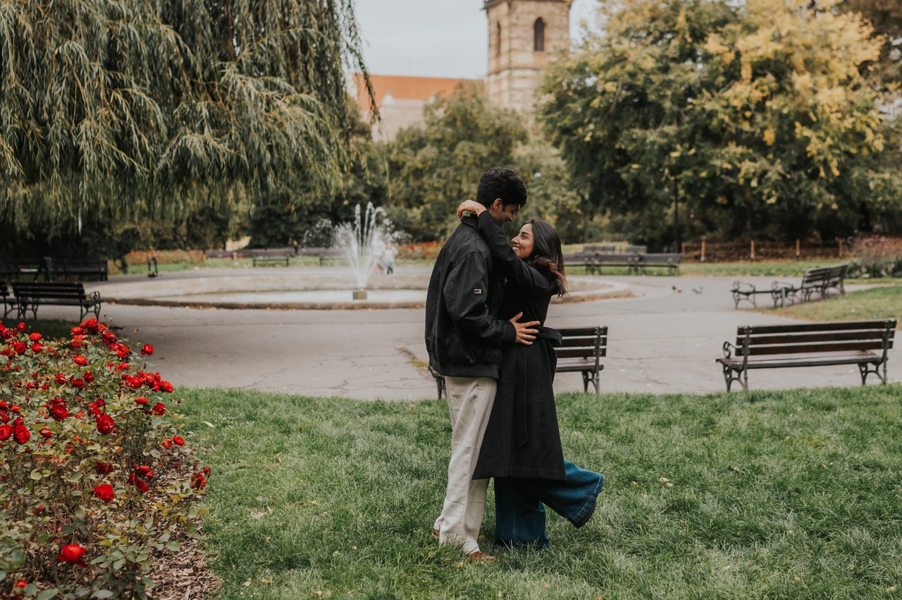 Prague couple photoshoot: a kiss among the Charles Square flowers Wide portrait of the couple kissing beside a flowerbed with the fountain behind them during their Prague couple photoshoot.