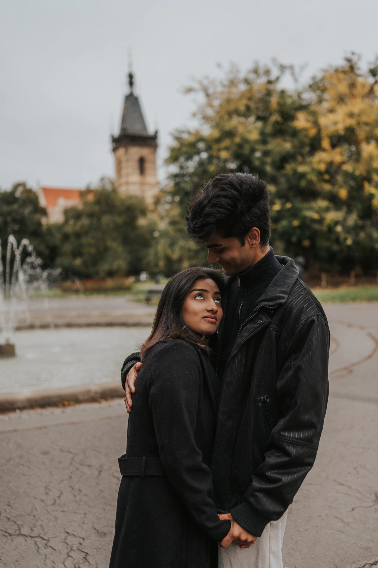 Couple embracing in front of the park fountain with the New Town Hall tower above them during their Prague couple photoshoot.