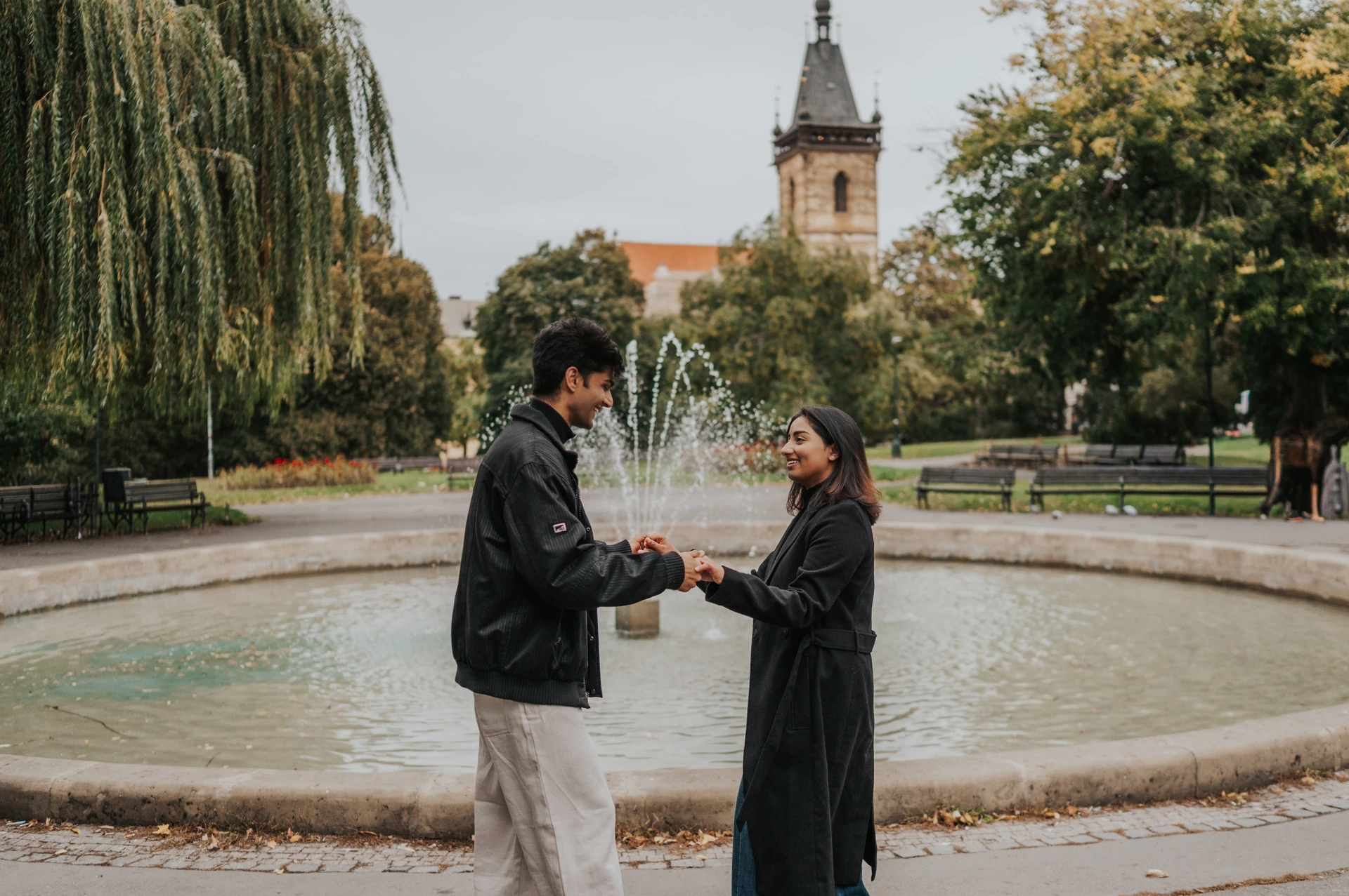 Prague couple photoshoot: holding hands by the fountain and New Town Hall tower Couple holding hands in front of a fountain with the New Town Hall tower above the trees during their Prague couple photoshoot.