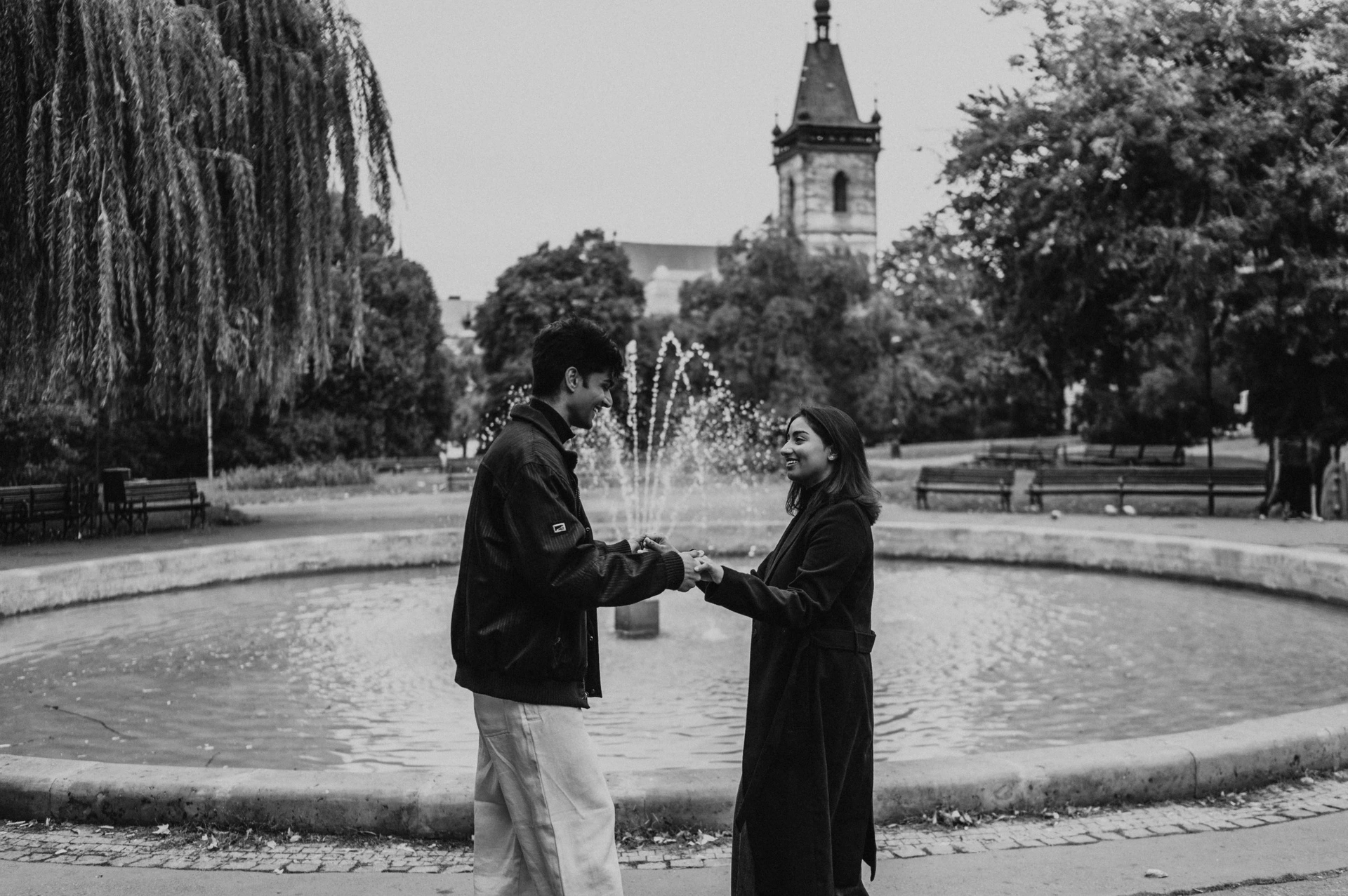 Black-and-white wide photograph of the couple holding hands beside the Charles Square Park fountain during their Prague couple photoshoot.