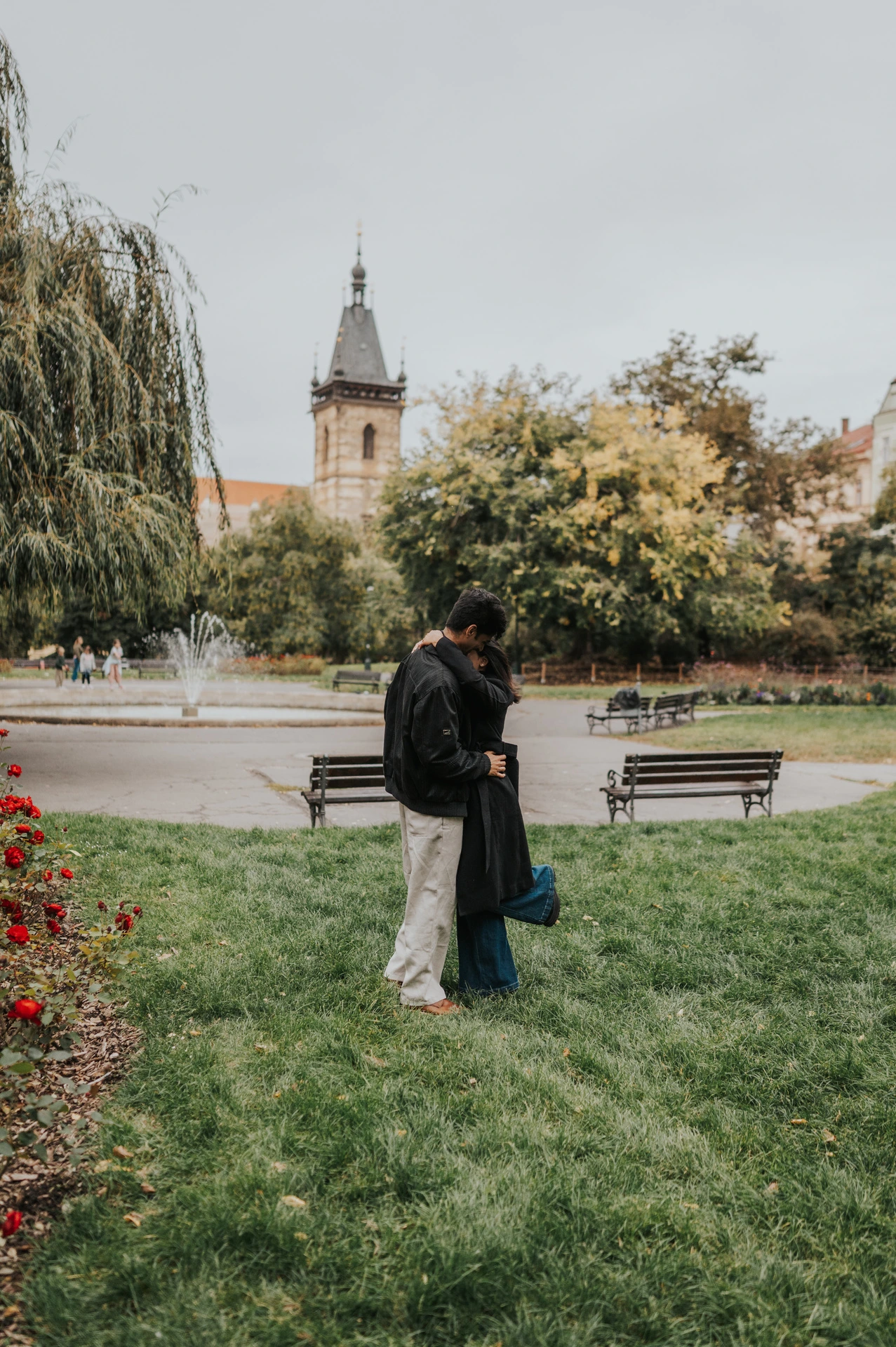 Couple embracing on the grass beside a flowerbed with the park fountain behind them during their Prague couple photoshoot.